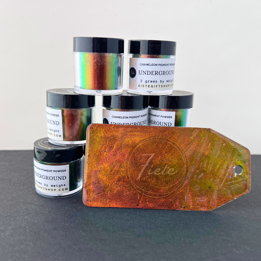 A display of Siete Gift Shop’s “Underground” chameleon pigment powder jars stacked in a small pyramid, showcasing their multicolor shift from green to copper. In front of the jars is a painted sample tag with a vivid green–gold color shift, placed on a black surface with a clean white background.