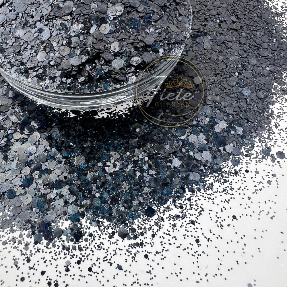 Dark charcoal and silver chunky holographic glitter mix with blue reflective sparkles spilled beside a clear container labeled Siete Gift Shop.