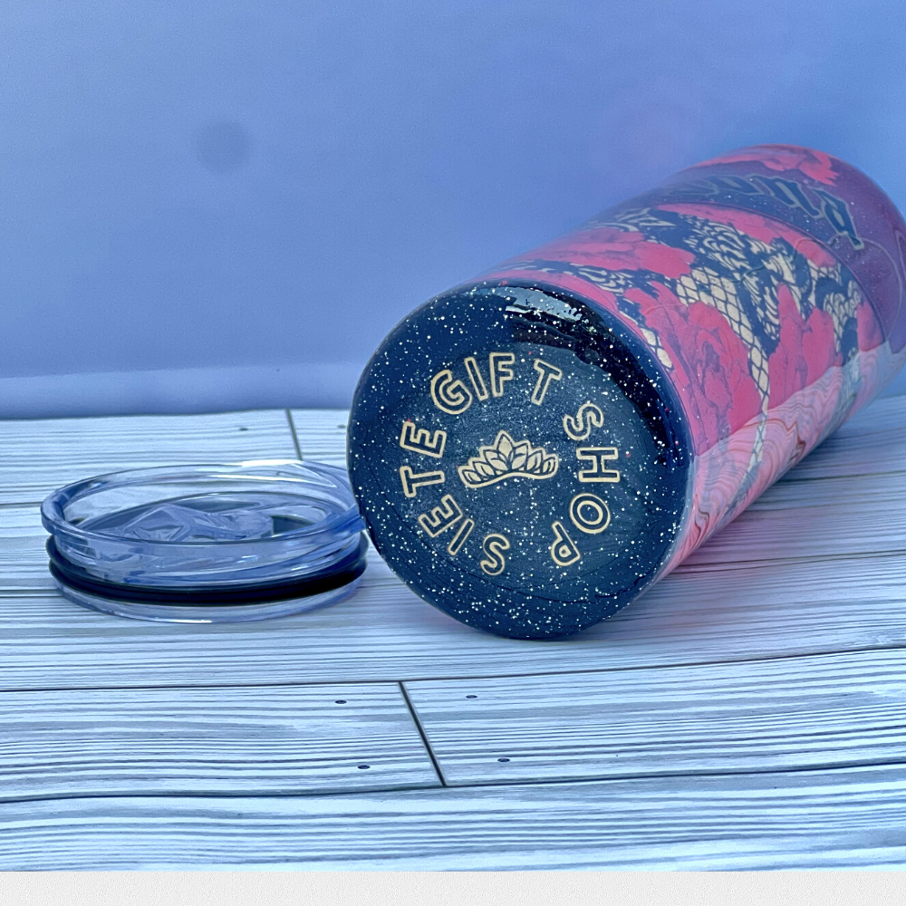 The tumbler lying flat to show the bottom of the cup which has matching black glitter. It has the words in a circular motion "Siete Gift Shop" in matching black and gold vinyl with a mini crown in the center.