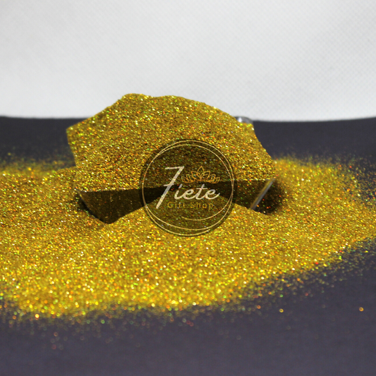 Extra fine glitter overflowing from a small clear container. The container is surrounded by the same glitters. This glitter is holographic and gold, reflecting light showing all the colors of the rainbow.