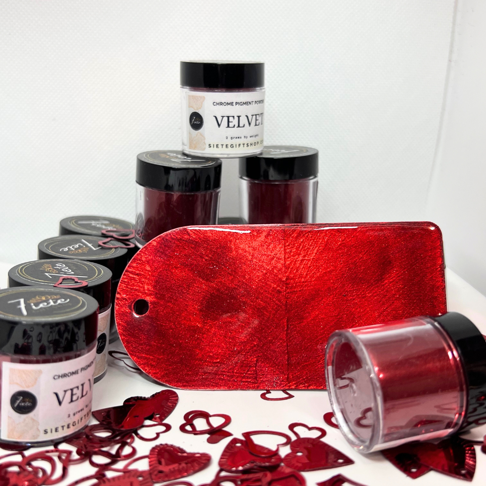 Several bottles of pigment powder on a white surface with white background, a metal gift tag that the red pigment has been applies to. red heart confetti surrounding the bottles.