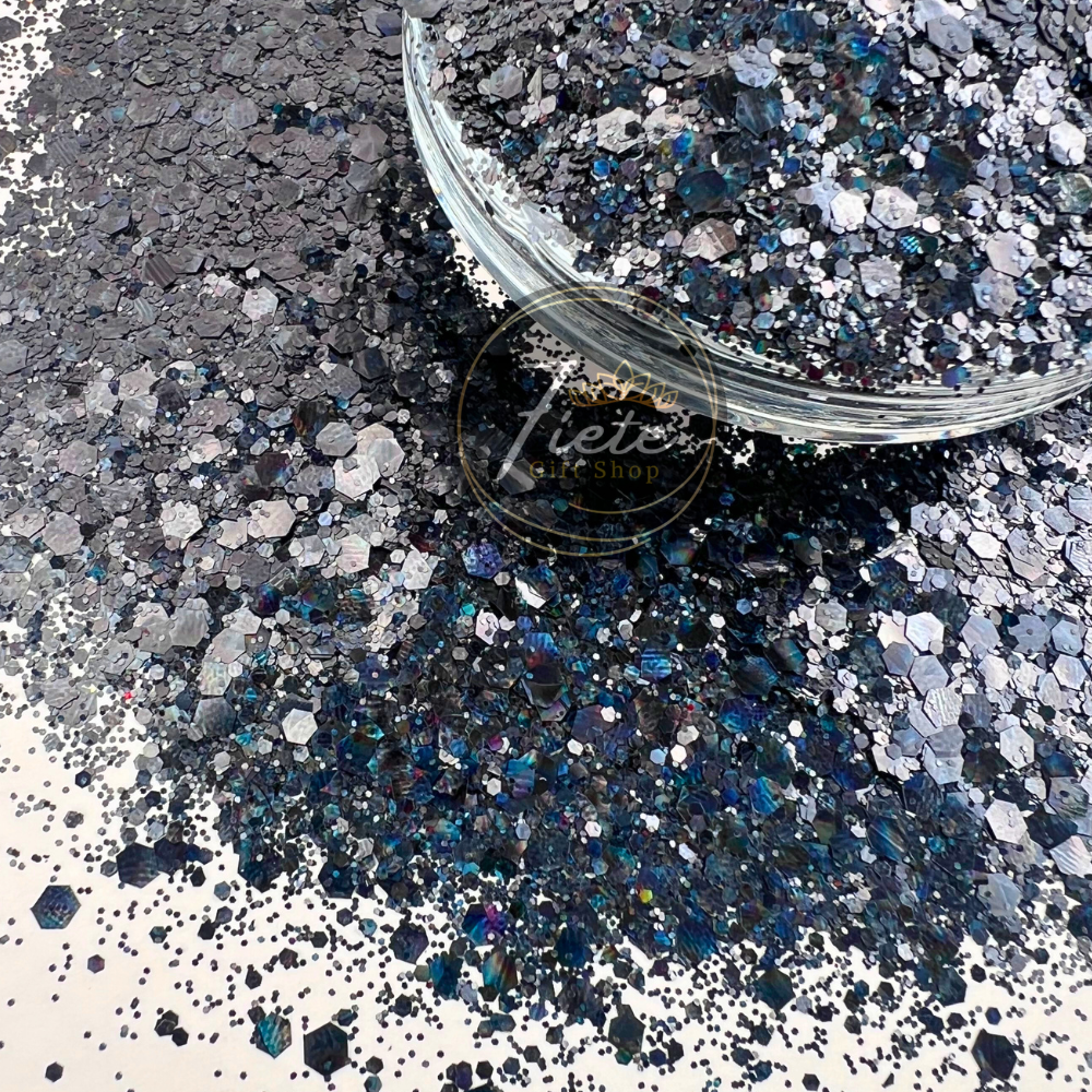 Dark charcoal and silver chunky holographic glitter mix with blue reflective sparkles spilled beside a clear container labeled Siete Gift Shop.