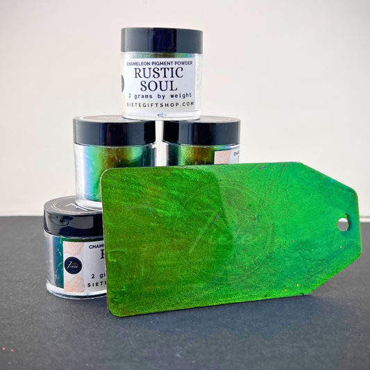 A stack of Siete Gift Shop’s “Rustic Soul” chameleon pigment powder jars arranged in a small pyramid, each showing a green-to-bronze color shift. In front of the jars is a painted sample tag with a bright green shimmering finish, placed on a dark surface with a white background.
