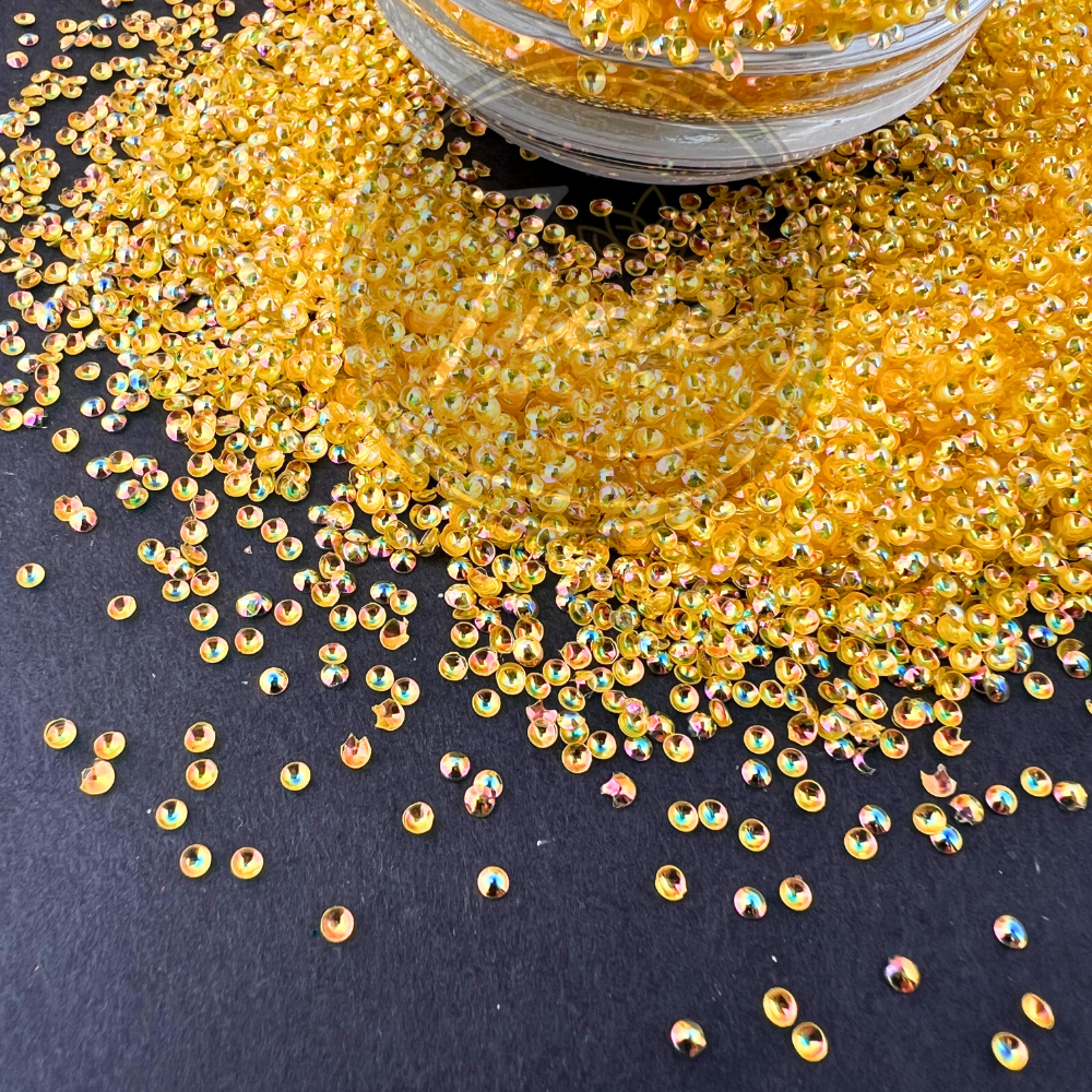 Close-up image of golden pearl-cut iridescent glitter named Belle, scattered across a dark surface with a jar overflowing, showing shimmering reflective colors in each piece.