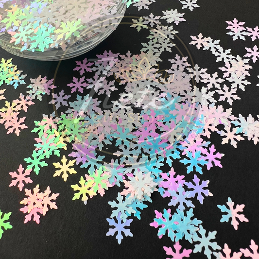 a collection of holographic, iridescent snowflake-shaped confetti scattered on a dark background. The snowflakes reflect different pastel shades—pinks, blues, greens, and purples—depending on the light.