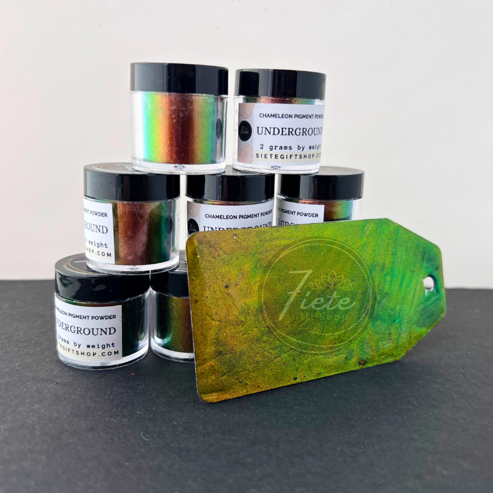 A display of Siete Gift Shop’s “Underground” chameleon pigment powder jars stacked in a small pyramid, showcasing their multicolor shift from green to copper. In front of the jars is a painted sample tag with a vivid green–gold color shift, placed on a black surface with a clean white background.