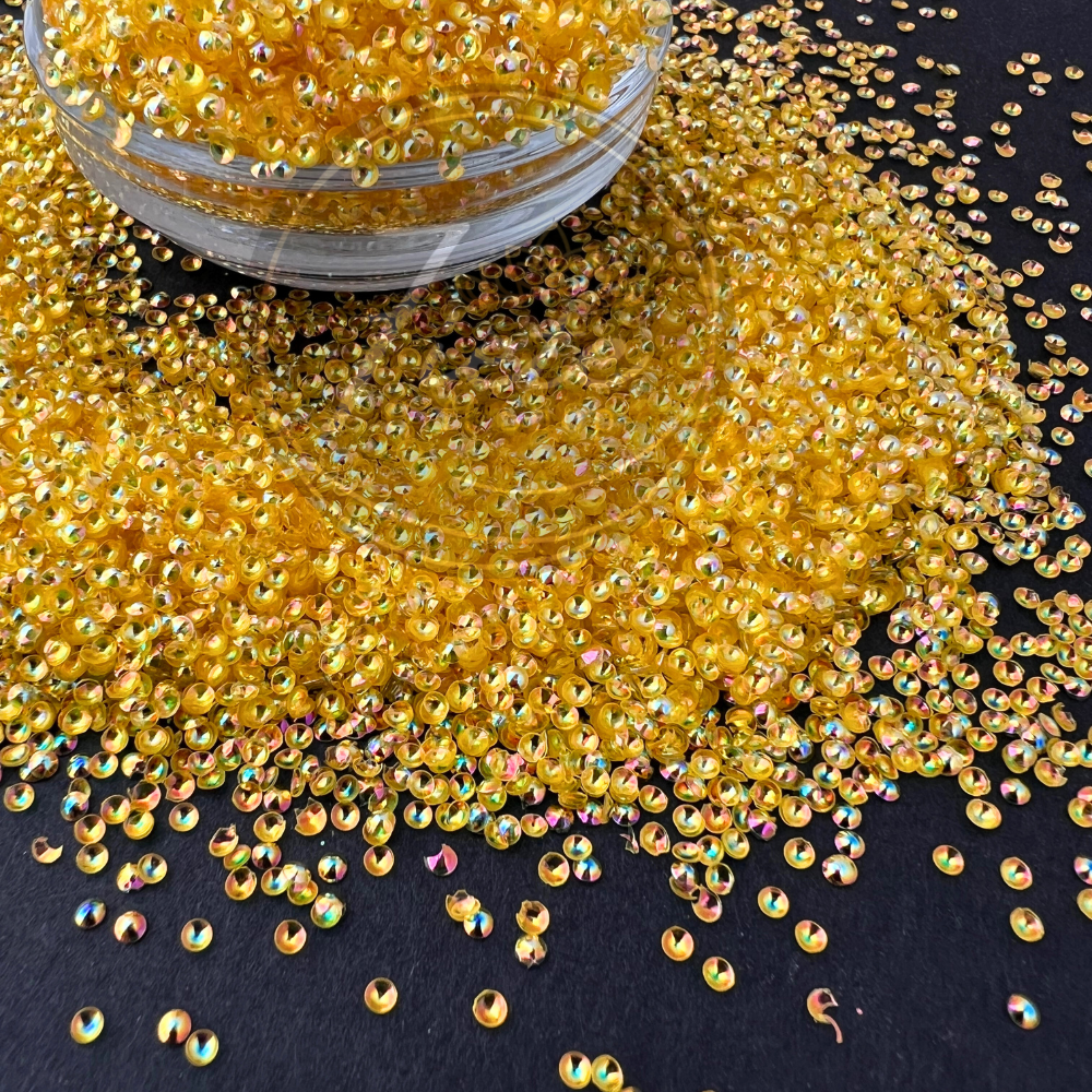 Close-up image of golden pearl-cut iridescent glitter named Belle, scattered across a dark surface with a jar overflowing, showing shimmering reflective colors in each piece.