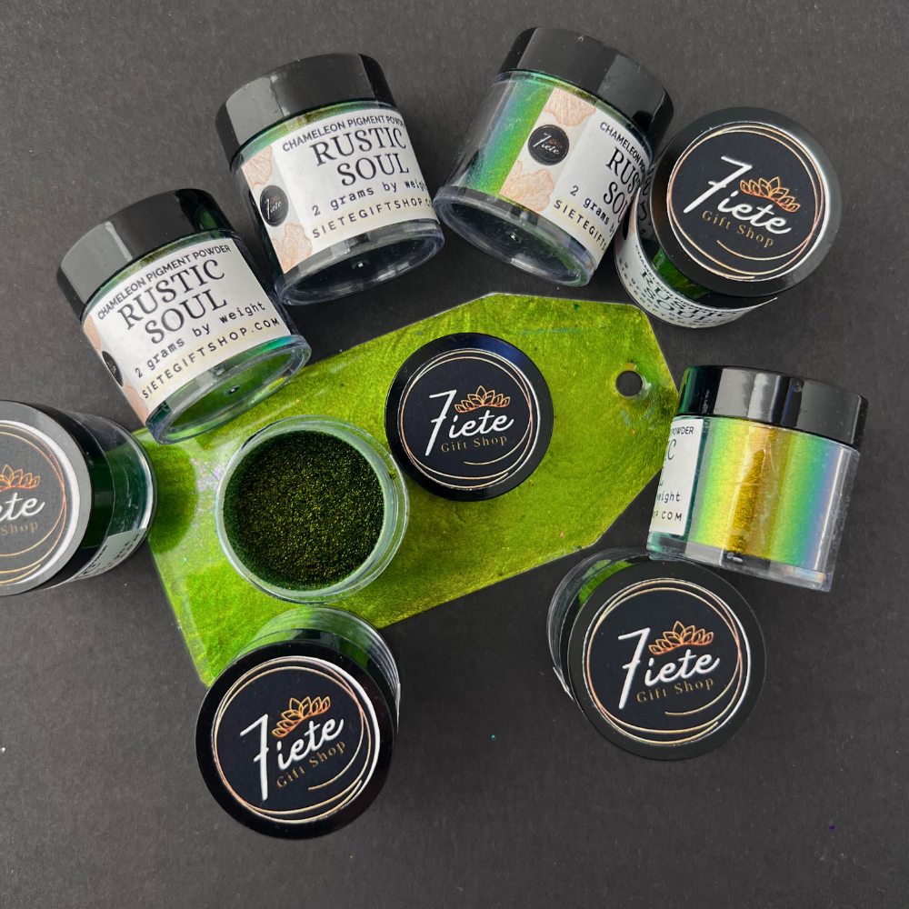 A stack of Siete Gift Shop’s “Rustic Soul” chameleon pigment powder jars arranged in a small pyramid, each showing a green-to-bronze color shift. In front of the jars is a painted sample tag with a bright green shimmering finish, placed on a dark surface with a white background.